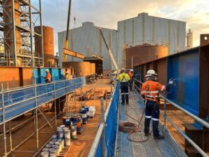 Industriële schilders in veiligheidskledij werken met verfspuiten op staalconstructies op bouwplaats met hijskranen